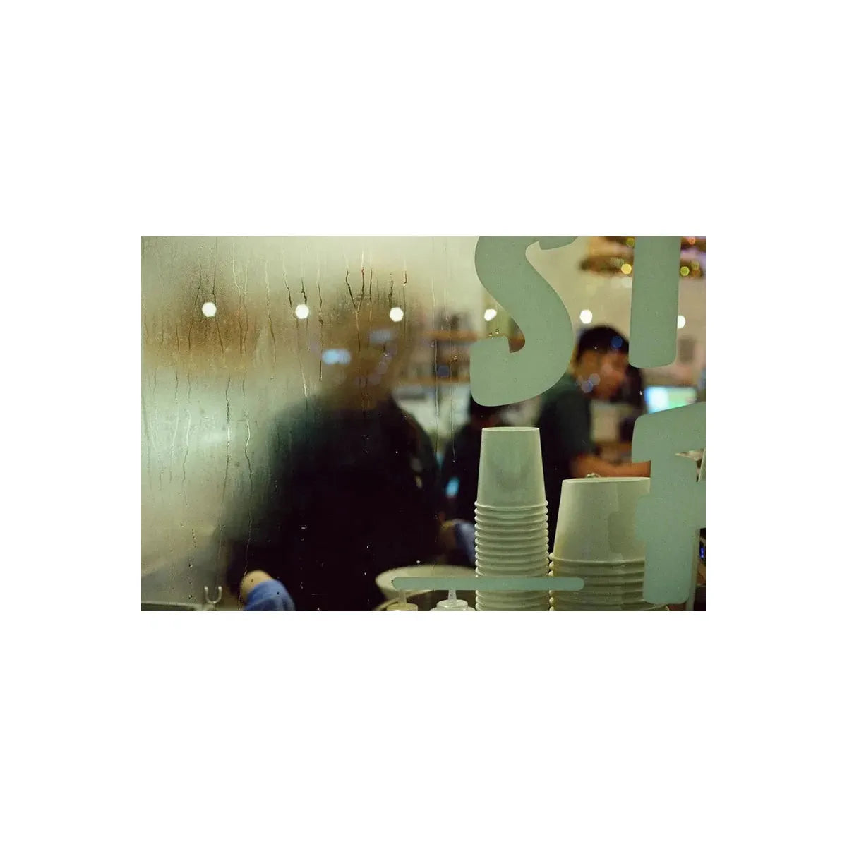 A close-up view through a wet, condensation-covered glass window, showing stacks of white disposable paper cups in the foreground and blurred figures of people inside a cafe or restaurant in the background.