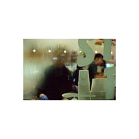 A close-up view through a wet, condensation-covered glass window, showing stacks of white disposable paper cups in the foreground and blurred figures of people inside a cafe or restaurant in the background.