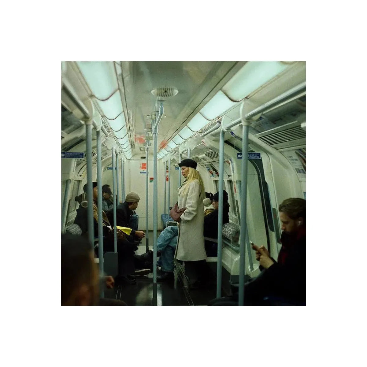 A person in a long white coat and black hat standing inside a subway train carriage, with other passengers seated around them.