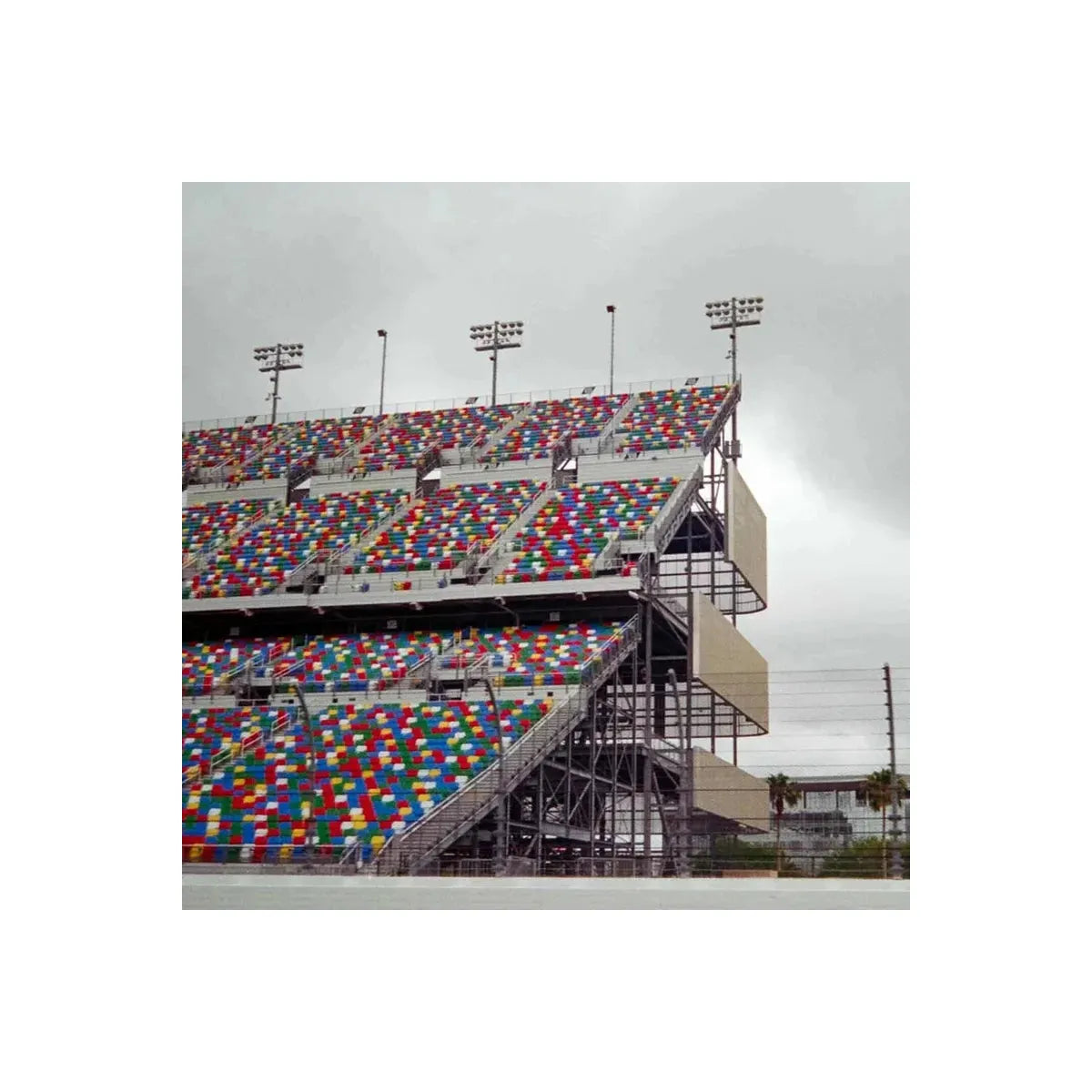 A large outdoor stadium with tiered seating covered in dense multicolored square tiles, photographed under an overcast gray sky. The structure includes metal support beams, track lighting, and a paved foreground in the foreground.