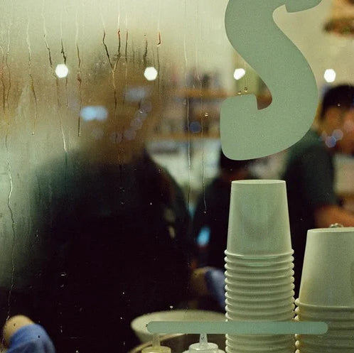 A close-up view through a wet, condensation-covered glass window, showing stacks of white disposable paper cups in the foreground and blurred figures of people inside a cafe or restaurant in the background.