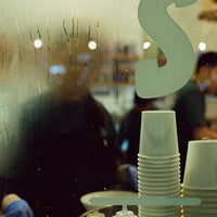 A close-up view through a wet, condensation-covered glass window, showing stacks of white disposable paper cups in the foreground and blurred figures of people inside a cafe or restaurant in the background.