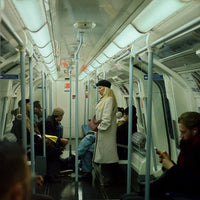 A person in a long white coat and black hat standing inside a subway train carriage, with other passengers seated around them.