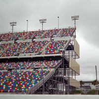 A large outdoor stadium with tiered seating covered in dense multicolored square tiles, photographed under an overcast gray sky. The structure includes metal support beams, track lighting, and a paved foreground in the foreground.