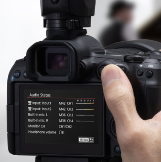 Close-up of a black DSLR camera body with a hand turning a dial, showing the rear LCD screen with audio status settings and a blurred person in the background.