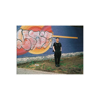 A person wearing a black t-shirt and black overalls stands in front of a blue brick wall with a large graffiti mural in red, orange, and yellow.