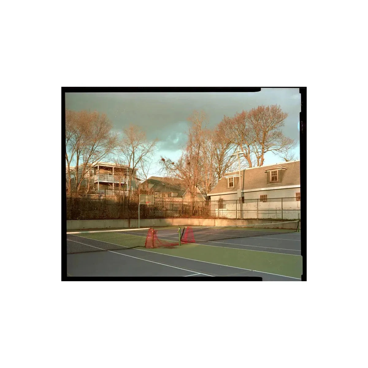 A vintage-style photograph of an outdoor tennis court with green and blue surfaces, orange cones, a fence, bare trees, and residential houses under a cloudy sky.