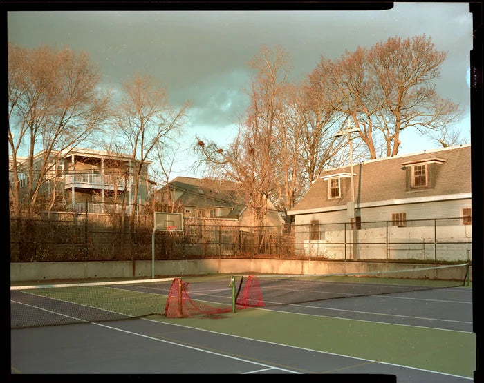 A vintage-style photograph of an outdoor tennis court with green and blue surfaces, orange cones, a fence, bare trees, and residential houses under a cloudy sky.