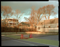 A vintage-style photograph of an outdoor tennis court with green and blue surfaces, orange cones, a fence, bare trees, and residential houses under a cloudy sky.