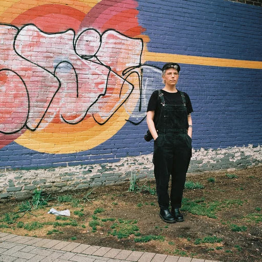A person wearing a black t-shirt and black overalls stands in front of a blue brick wall with a large graffiti mural in red, orange, and yellow.
