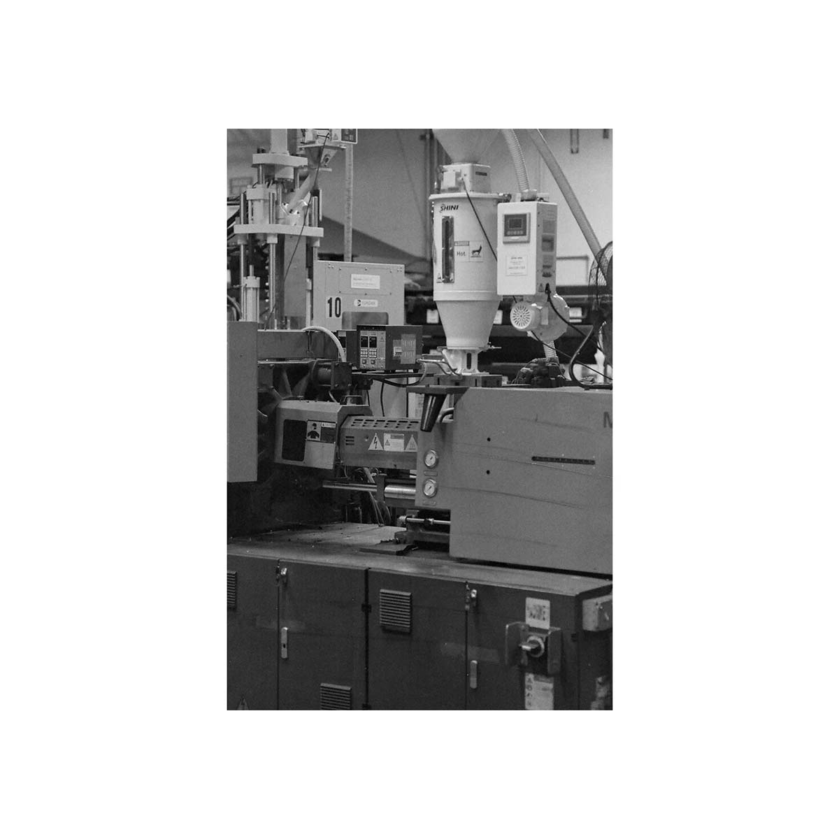 A black and white photograph of an industrial injection molding machine in a factory setting, featuring a hopper, control panel, and metal components.
