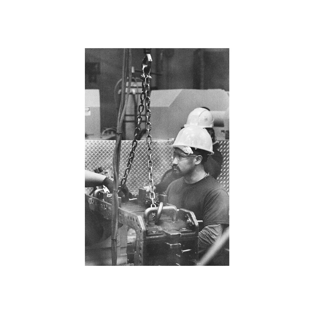 A black and white photograph depicting a worker in a hard hat and safety glasses operating heavy machinery suspended by chains in an industrial setting.