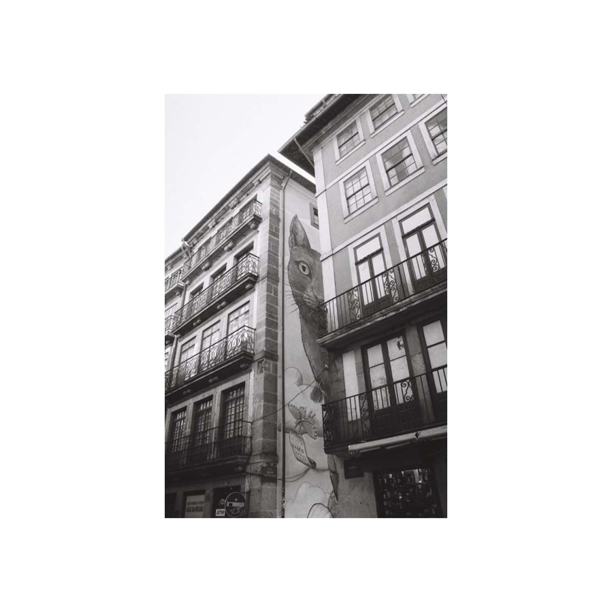 A black and white photograph of old European-style buildings with balconies and a large mural or painted figure on the side of one building, centered within a white border.