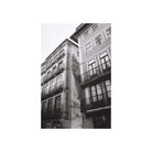 A black and white photograph of old European-style buildings with balconies and a large mural or painted figure on the side of one building, centered within a white border.