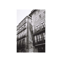A black and white photograph of old European-style buildings with balconies and a large mural or painted figure on the side of one building, centered within a white border.