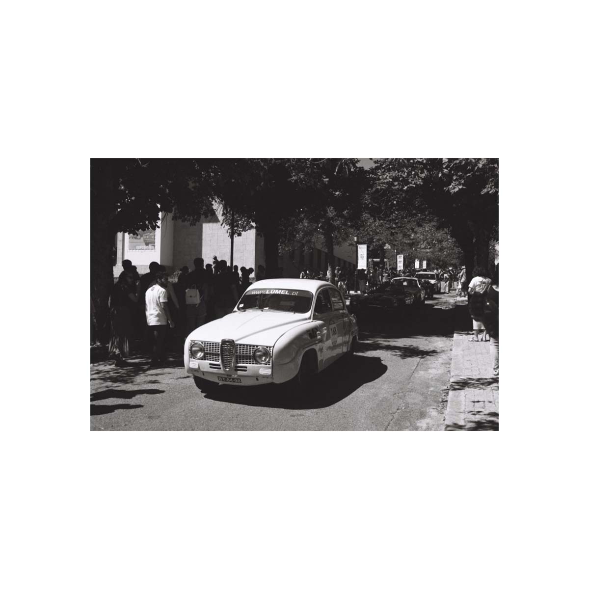 A black and white photograph print featuring a vintage car driving down a tree-lined street with onlookers, centered on a white background.