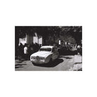 A black and white photograph print featuring a vintage car driving down a tree-lined street with onlookers, centered on a white background.