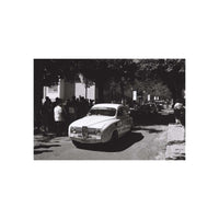 A black and white photograph print featuring a vintage car driving down a tree-lined street with onlookers, centered on a white background.