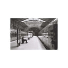 A black and white photograph of a train station platform, showing two trains at the tracks, a seated person on a bench, and an arched station roof overhead.