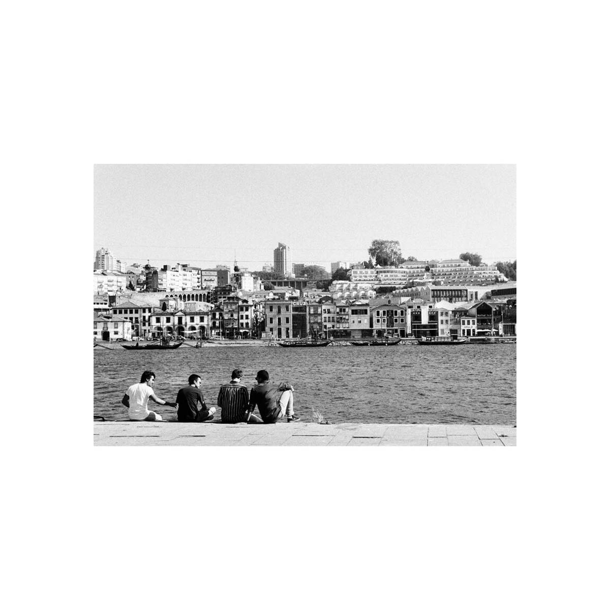 A black and white photographic print depicting a waterfront scene with several people sitting on a ledge in the foreground, looking out over a body of water toward a dense city skyline with buildings and boats under a clear sky.