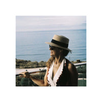 A woman wearing a straw boater hat with a black band, sunglasses, and a white top with a scalloped red edge, standing outdoors with the ocean in the background.