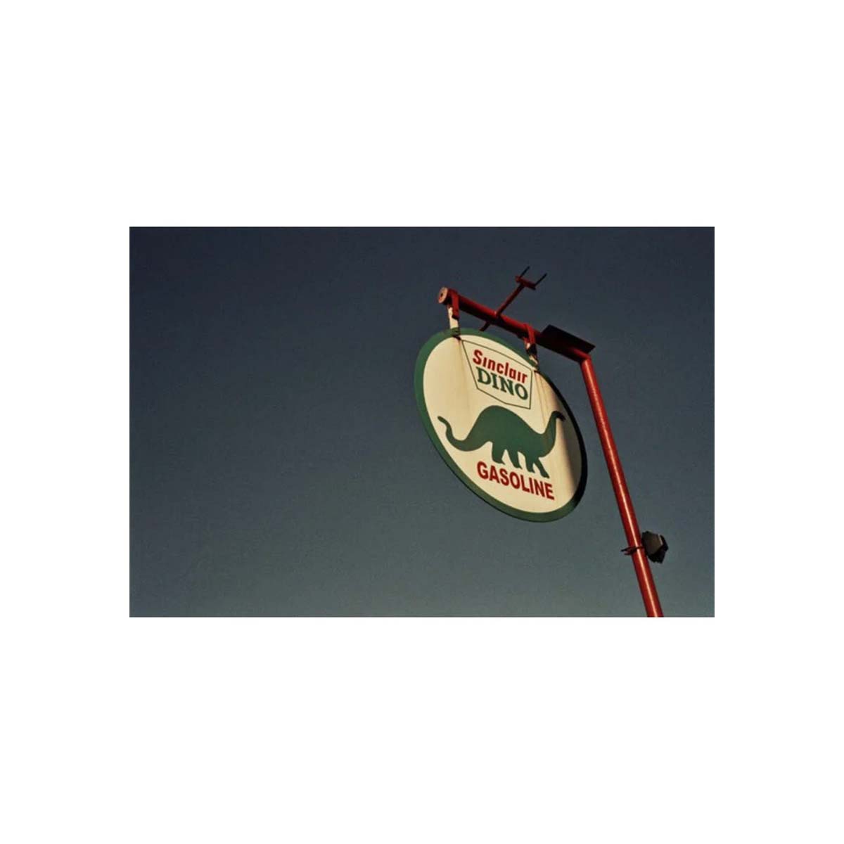 A vintage-style round sign for 'Suncor DOW Gasoline' hanging from a red metal pole against a dark blue sky. The sign has a cream background with a green dinosaur silhouette and red lettering.