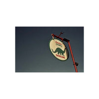 A vintage-style round sign for 'Suncor DOW Gasoline' hanging from a red metal pole against a dark blue sky. The sign has a cream background with a green dinosaur silhouette and red lettering.