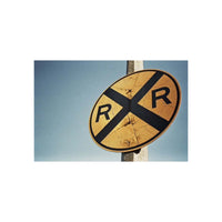 A vintage oval railroad crossing sign with a black cross and the letters 'R' and 'R' on a yellow background, mounted on a pole against a blue sky.