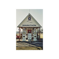 A vintage-style gas station building with two white gas pumps under a gabled roof, shown against a pale sky. The building has large 'MARATHON' lettering on the front, with smaller signage including 'MOM & POP'S' and '1947'.