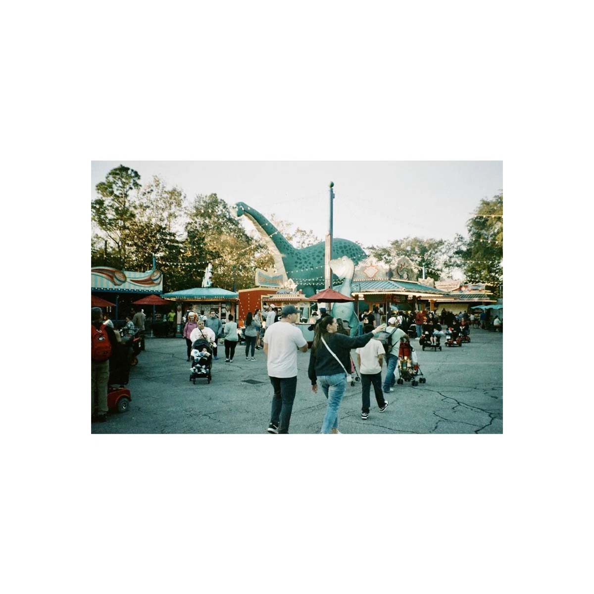A square photographic print of a crowded outdoor plaza with people walking toward a large dinosaur sculpture, surrounded by market stalls and trees under a pale sky.