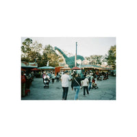 A square photographic print of a crowded outdoor plaza with people walking toward a large dinosaur sculpture, surrounded by market stalls and trees under a pale sky.