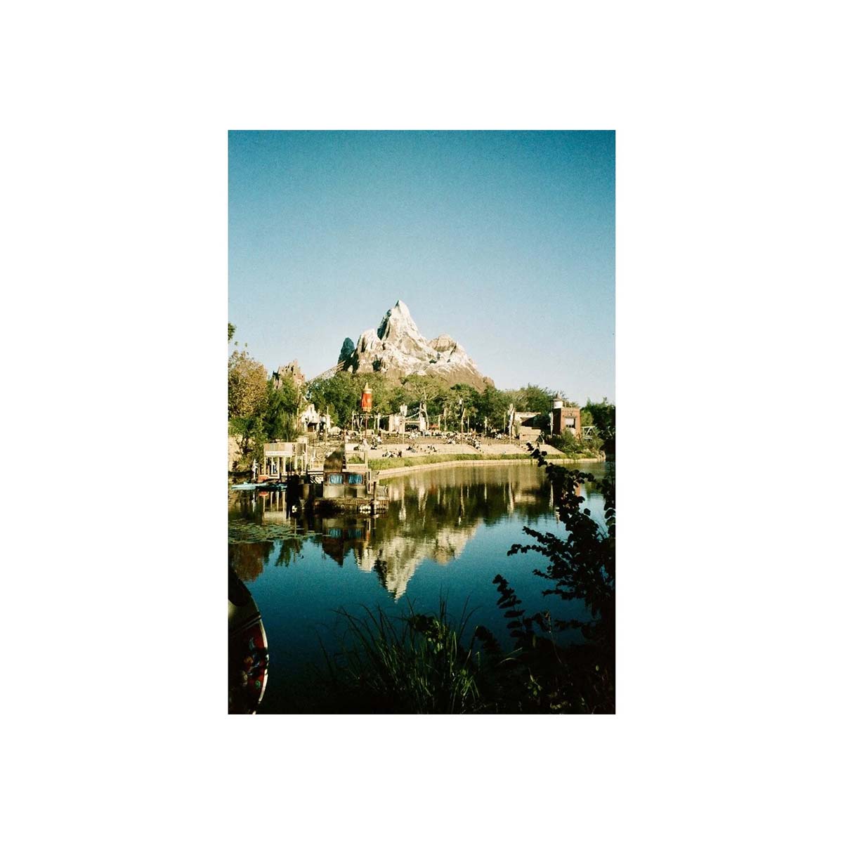 A vertical photographic print of a scenic landscape featuring a mountain peak reflected in a calm lake, with trees, buildings, and a clear blue sky. The print is centered on a wide white background.