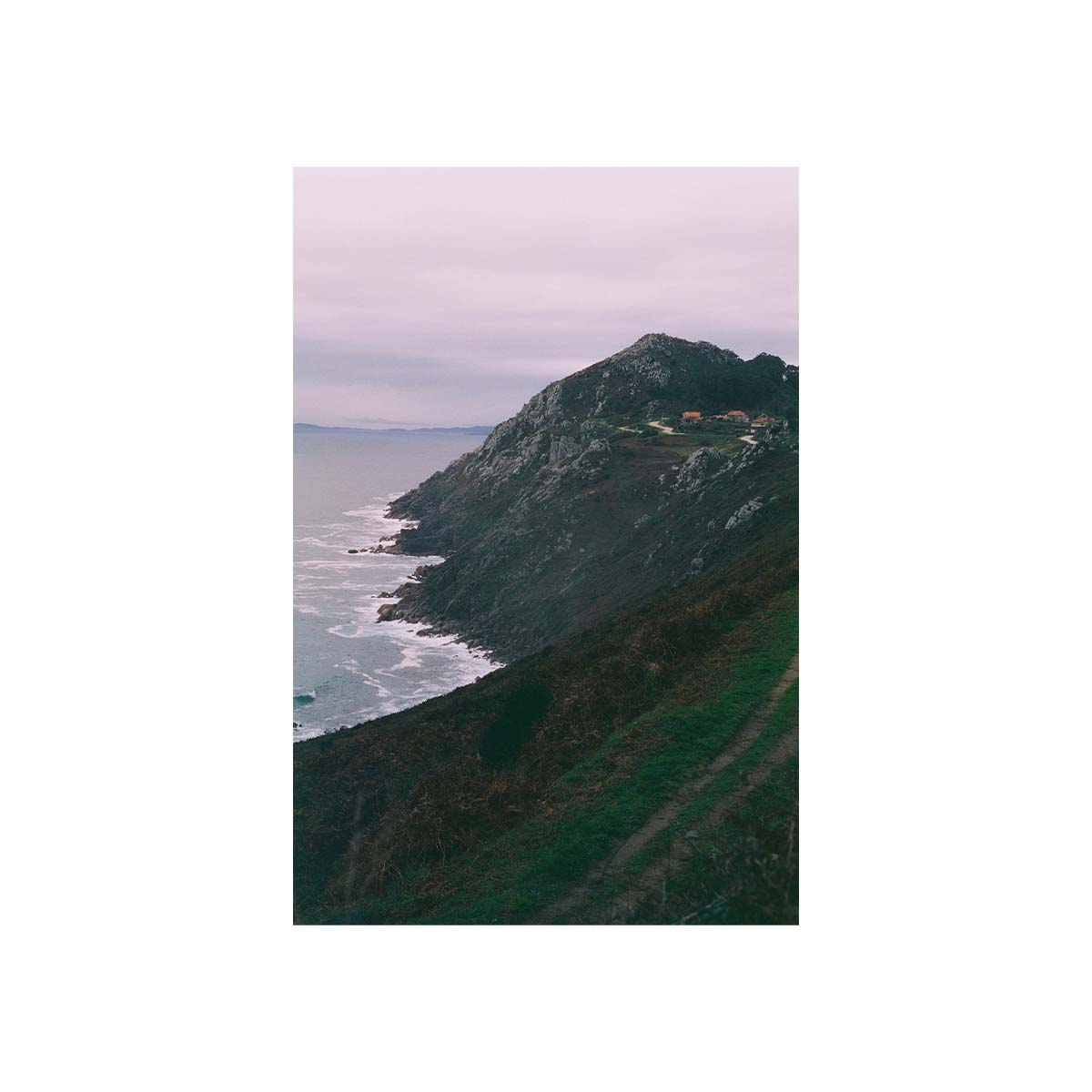 A vertical photographic print of a rugged coastal cliffside overlooking the ocean under a pale cloudy sky, with a small cluster of buildings near the peak.