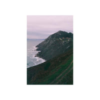 A vertical photographic print of a rugged coastal cliffside overlooking the ocean under a pale cloudy sky, with a small cluster of buildings near the peak.