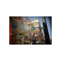 A square art print or photograph depicting a colorful urban alleyway with hanging multicolored flags, a mural on a building, and brick apartment buildings under a blue sky.