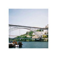 A scenic photograph of a large arched bridge spanning a river, with buildings and green hills in the background and boats in the foreground, presented as a square print centered on a white background.
