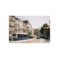 A vintage-style photograph print depicting a European street scene with a blue and white bus, multi-story buildings, and pedestrians on a sidewalk.