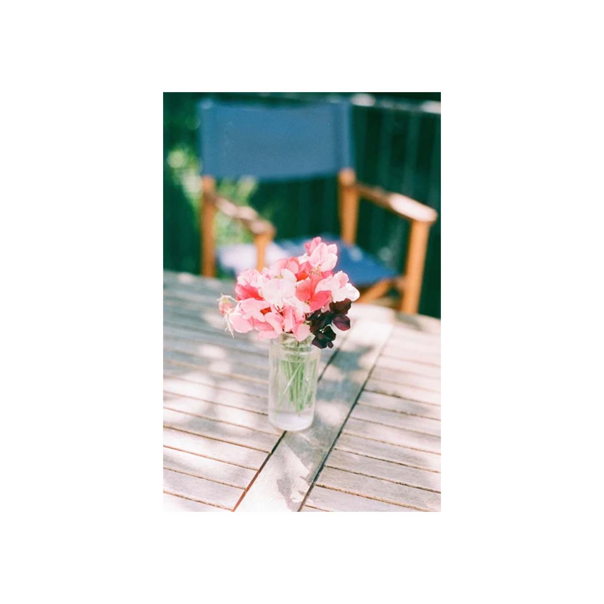 A small bouquet of pink flowers in a clear glass jar vase, placed on a weathered wooden table with a blue chair and green fence blurred in the background.