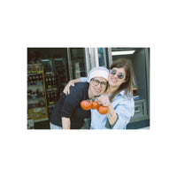 Two women posing together in front of a store entrance, holding a cluster of red tomatoes. One woman wears a white head covering and glasses, while the other wears a light blue shirt and sunglasses.