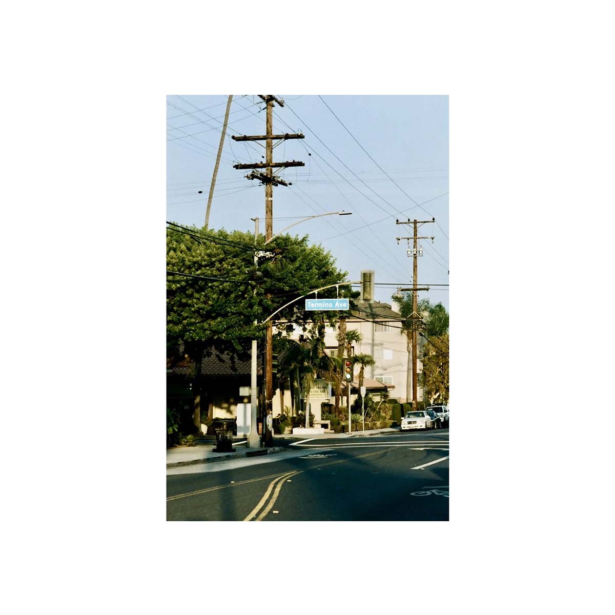 A vertical photographic print of a quiet residential street corner, featuring tall wooden utility poles with multiple cross-arms, overhead wires, green trees, a small building, and a few cars on the road under a pale blue sky.