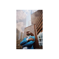 Man in a blue blazer and white turtleneck posing against a metal pillar in a city with tall glass buildings.