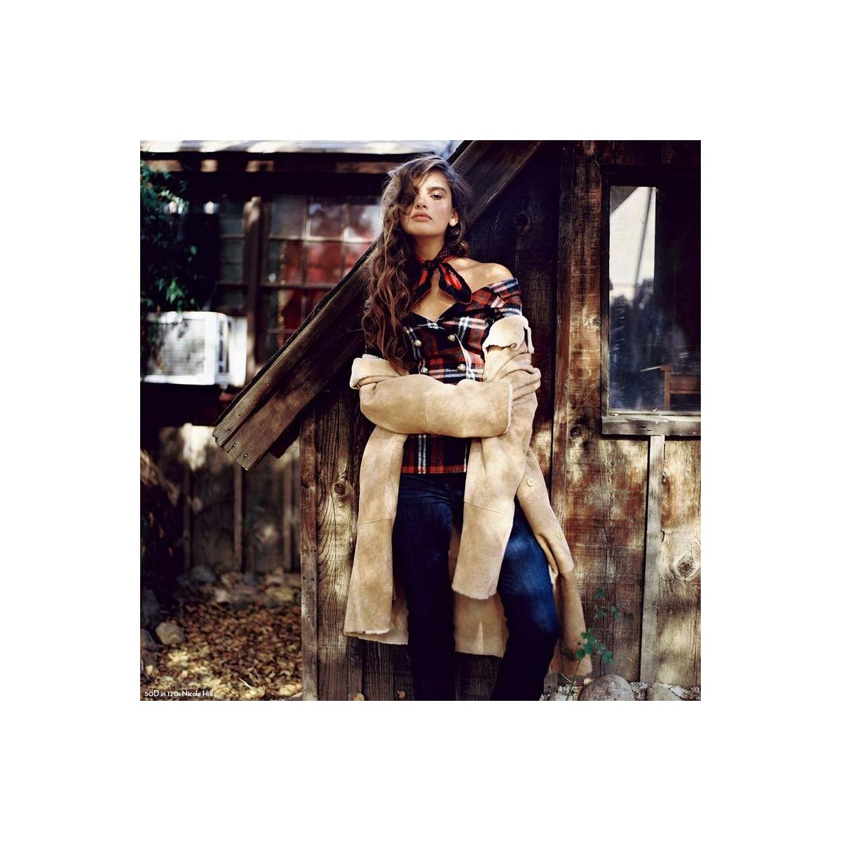 Woman in a plaid top and tan coat leaning against a rustic wooden shed, captured on CineStill 50Daylight 120 film.