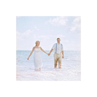 Bride and groom in wedding attire walking hand-in-hand through bright, sunlit ocean water on a beach.