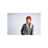 A woman wearing a bright red knit beanie, an olive green jacket, and a black-and-white checkered shirt, standing against a light gray studio background.