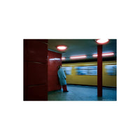 Person in a light blue coat leaning against a red subway wall as a blurred yellow train passes by in low light.