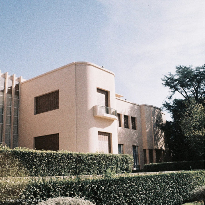 A modernist-style stucco building in a pale peach-pink color, featuring angled corners, dark window frames, a small balcony, and a glass curtain wall on the left, set behind a green hedge under a blue sky.