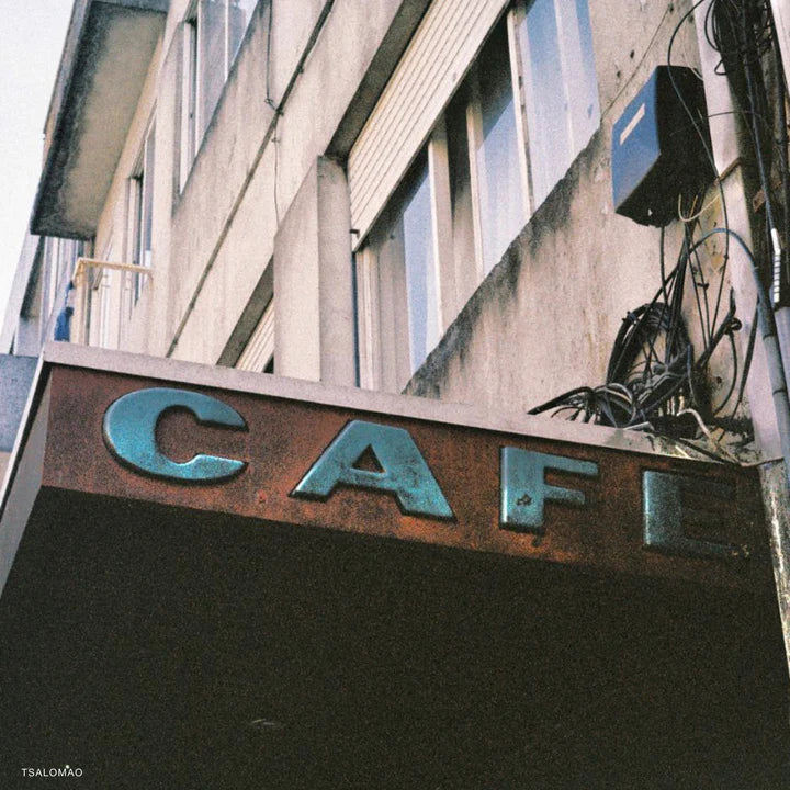 A close-up, low-angle photograph of a weathered metal sign spelling 'CAFE' in distressed teal-blue letters mounted on a brown awning, set against the facade of a concrete building with windows and exposed wiring.