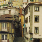 A narrow urban alleyway between closely packed apartment buildings with cream, beige, and ochre stucco walls, brown tiled roofs, stone trim, balconies, and a staircase leading down the center.