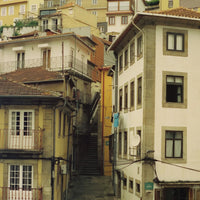 A narrow urban alleyway between closely packed apartment buildings with cream, beige, and ochre stucco walls, brown tiled roofs, stone trim, balconies, and a staircase leading down the center.