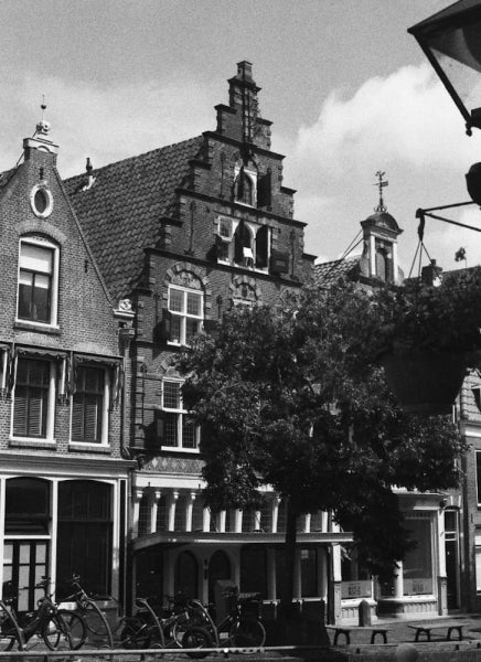 Black and white photograph of traditional European townhouses with stepped gables, bicycles parked along the street, and a large tree in the foreground.
